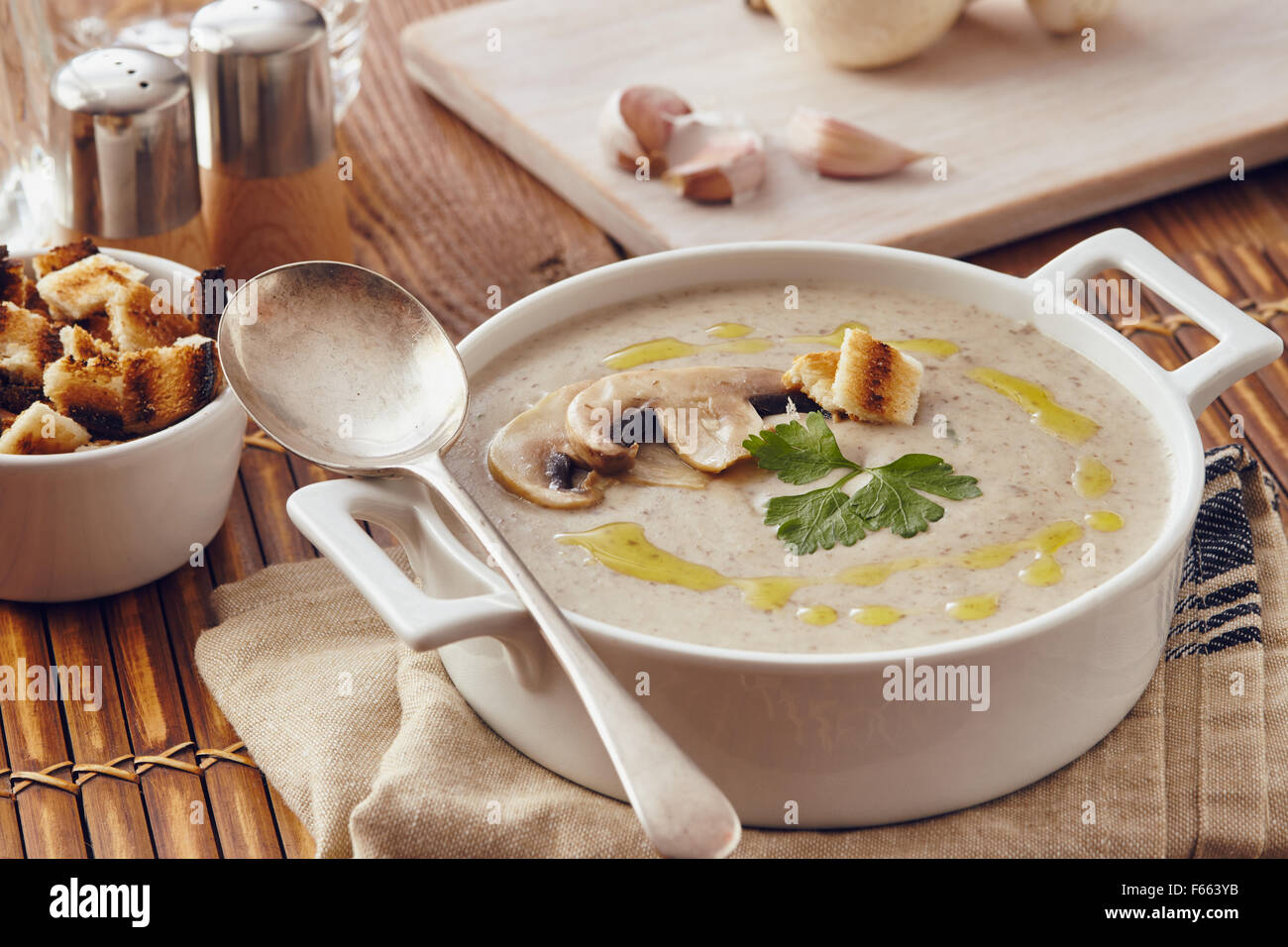 Champignon Cremesuppe auf einem Tisch mit Brot, Knoblauch und rohe Pilze auf einem Schneidebrett weiß. Salz und Pfeffer auf dem Tisch. Stockfoto
