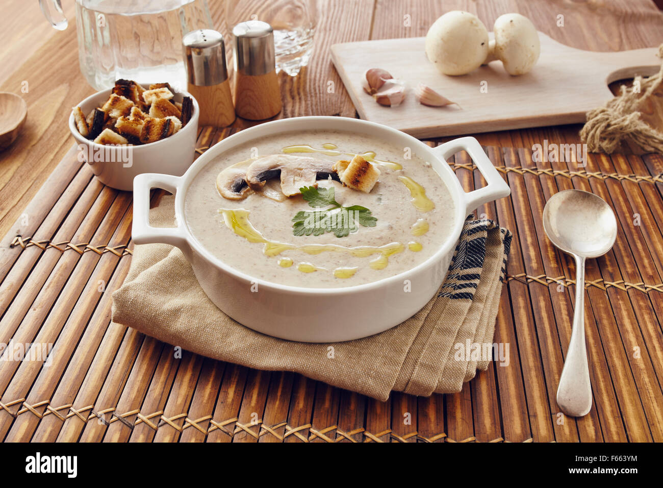 Champignon Cremesuppe auf einem Tisch mit Brot, Knoblauch und rohe Pilze auf einem Schneidebrett weiß. Salz und Pfeffer auf dem Tisch. Stockfoto