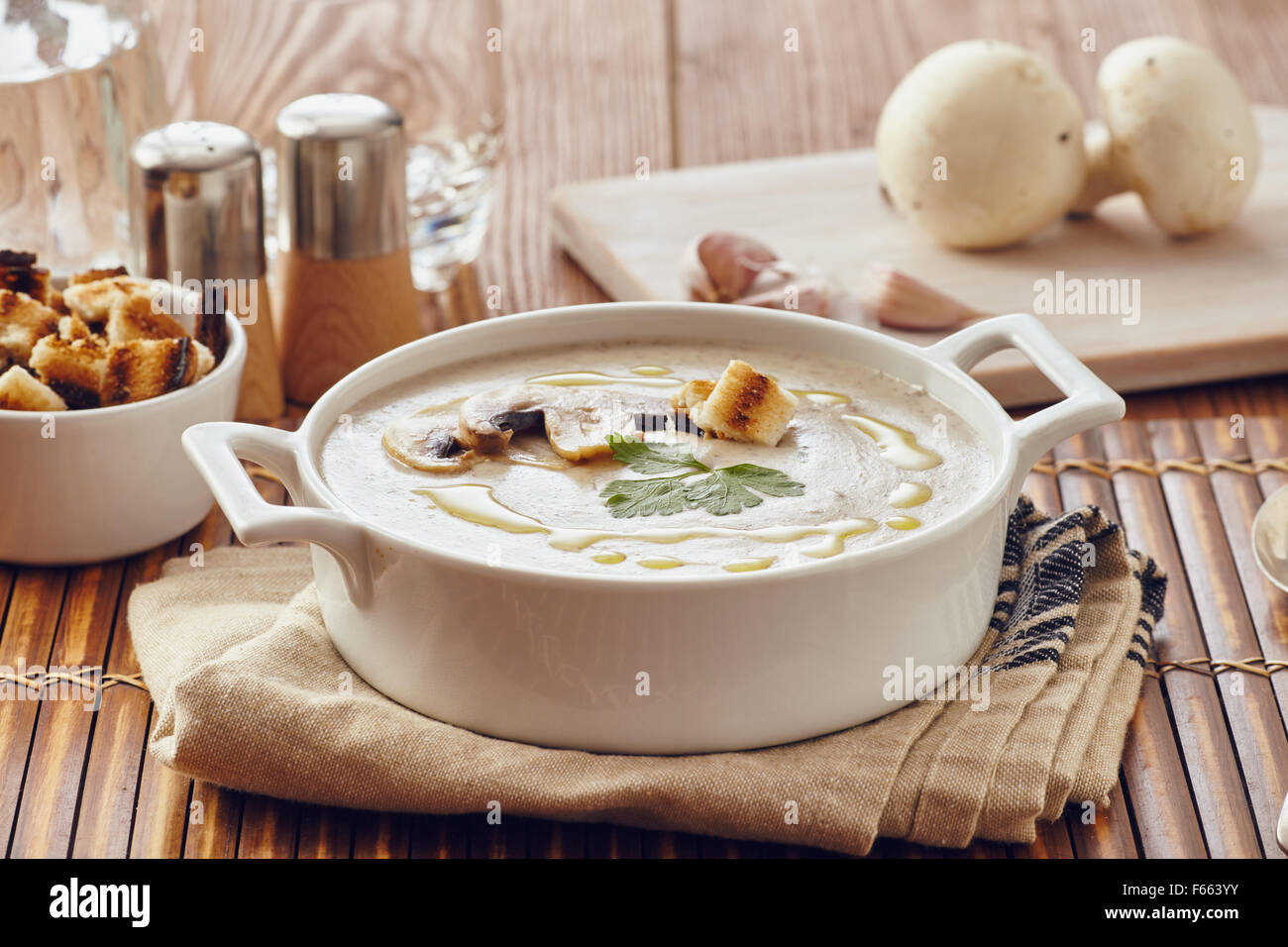 Champignon Cremesuppe auf einem Tisch mit Brot, Knoblauch und rohe Pilze auf einem Schneidebrett weiß. Salz und Pfeffer auf dem Tisch. Stockfoto