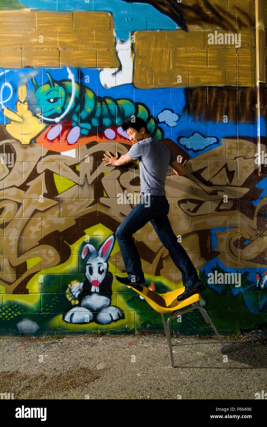 Asiatischer Mann stehend auf einem Stuhl gegen die Wand, dass graffitied wurde. ist ein Spaß und casual Bild, er treibt den Stuhl über. Stockfoto