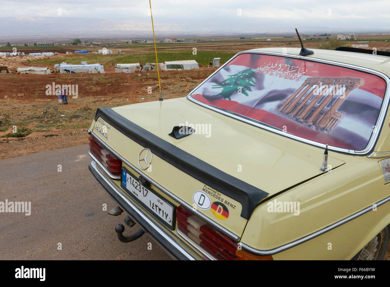 Libanon Beqaa Tal, Deir el Ahmad, Lager für syrische Flüchtlinge, alte deutsche Mercedes Benz 200 D Auto mit alten deutschen country code Kennzeichen D, libanesische Flagge und Bild von Baalbek Tempel, Hintergrund Anti Gebirge Libanon und Syrien Stockfoto