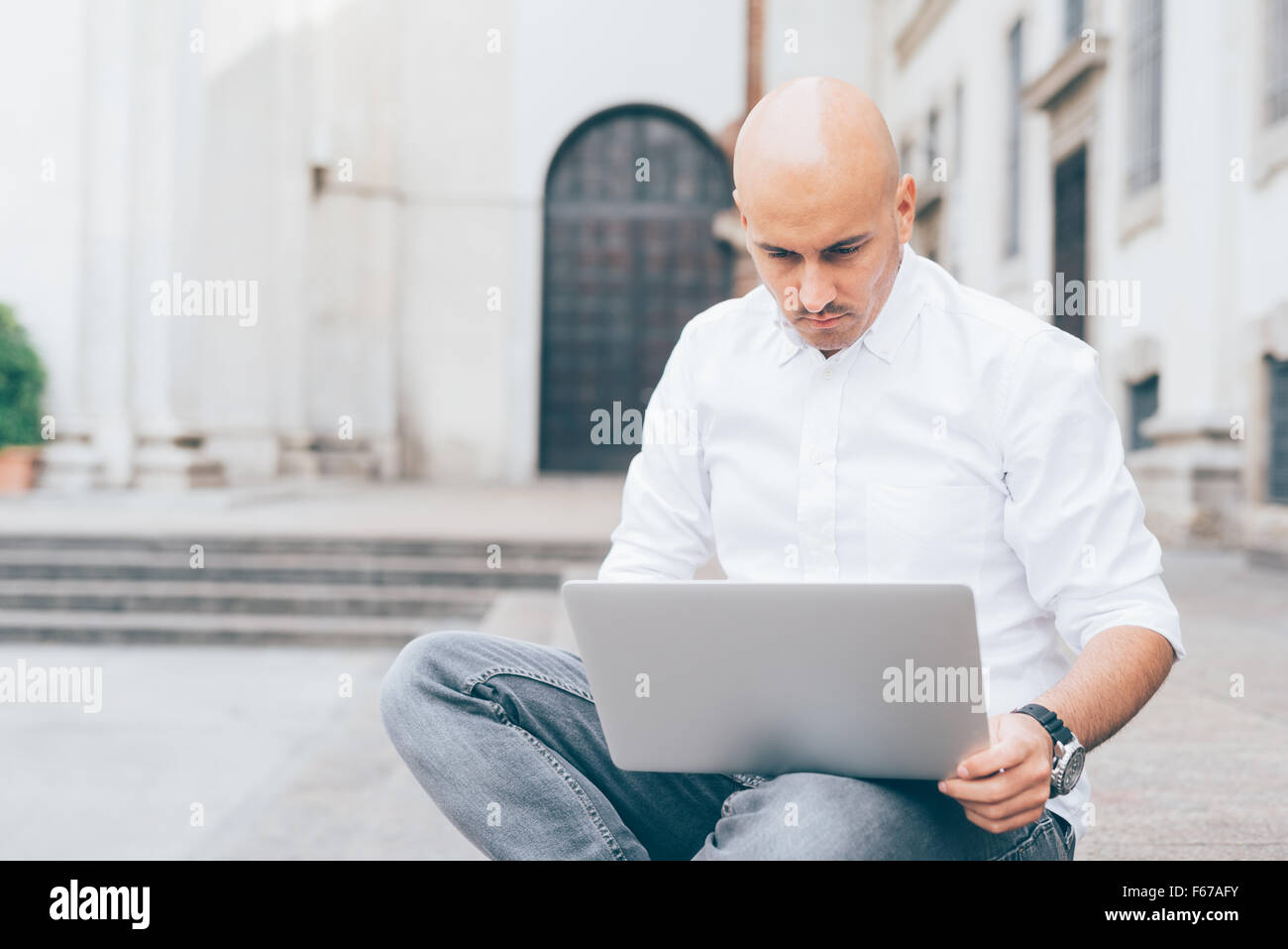 Junge hübsche kaukasischen kahl Geschäftsmann sitzt auf einer Treppe mit einem Laptop, stützte sich auf seinen Knien, auf der Suche nach unten den Bildschirm, nachdenklich - arbeiten, nachdenklich, beschäftigt Konzept Stockfoto