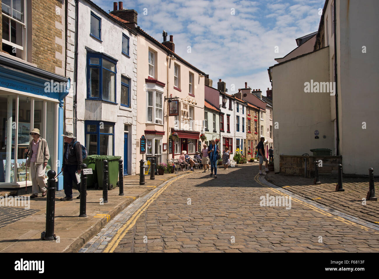 Die Menschen von einer Kneipe sitzen oder mach einen Sommer Spaziergang entlang der sonnigen gepflasterten Hauptstraße - malerischen Fischerdorf Dorf Staithes, North Yorkshire, England, UK. Stockfoto