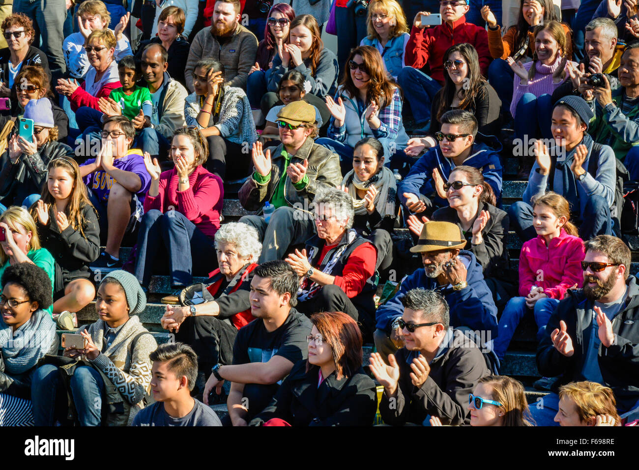 Eine große, multiethnische, vielfältige Gruppe von Menschen, die auf Treppen draußen in Reihen sitzen, als Publikum, das eine Live-Performance beobachtet und fröhlich reagiert Stockfoto