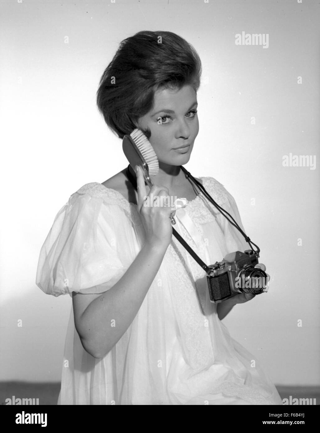Ein Schwarzweißfoto aus den 1960er Jahren, das ein weibliches Model in einem Studio zeigt, aufgenommen für eine Turners Photography-Werbekampagne. Das Bild zeigt das Modell in einer neutralen Pose, mit einem Fokus auf die Pentax-Kamera und die Mode der Epoche, die Modellierungstrends und fotografische Praktiken der Mitte des 20. Jahrhunderts veranschaulicht. Stockfoto