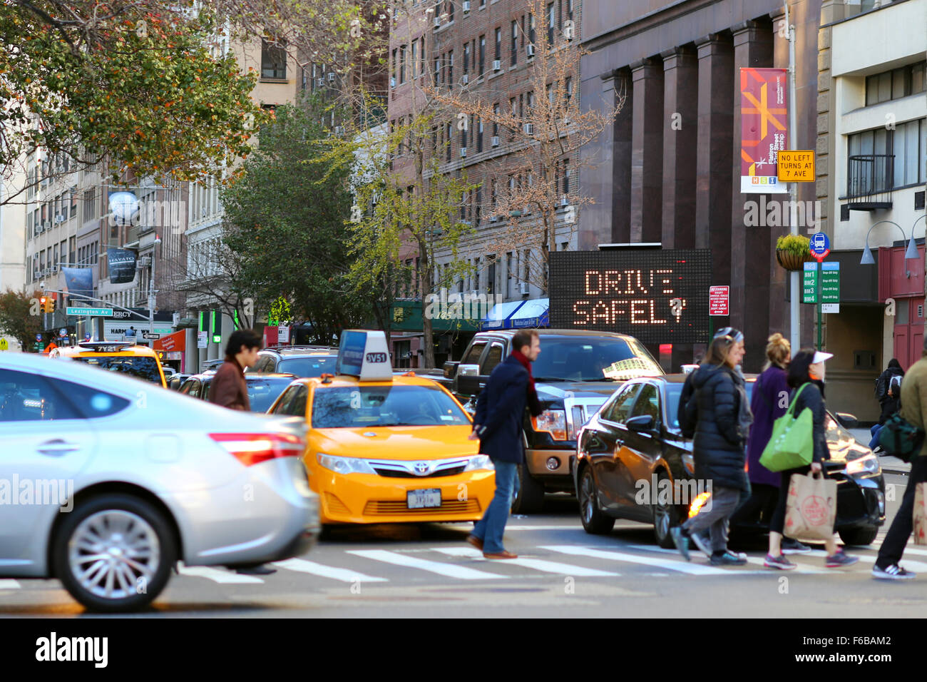 Ein elektronisches Schild blinkt Fahren Sie sicher in der Nähe einer Kreuzung von New York City, während Fußgänger über eine Straße neben Autos gehen, die auf dem Fußgängerübergang angehalten wurden. Stockfoto