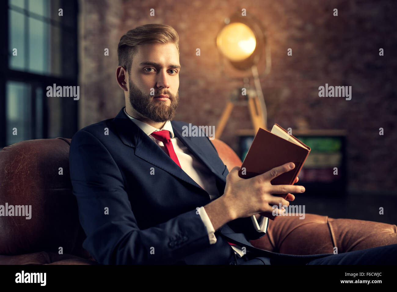 Junge hübsche Geschäftsmann mit Bart im schwarzen Anzug sitzen auf Stuhl Buch zu lesen. Stockfoto