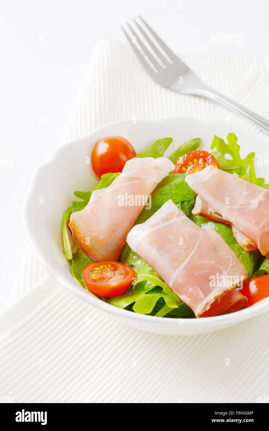 Schüssel mit getrockneten Schweinefleisch Schinken, Rucola und Spinat Blätter und halbierte Kirschtomaten auf weißen Tischset Stockfoto