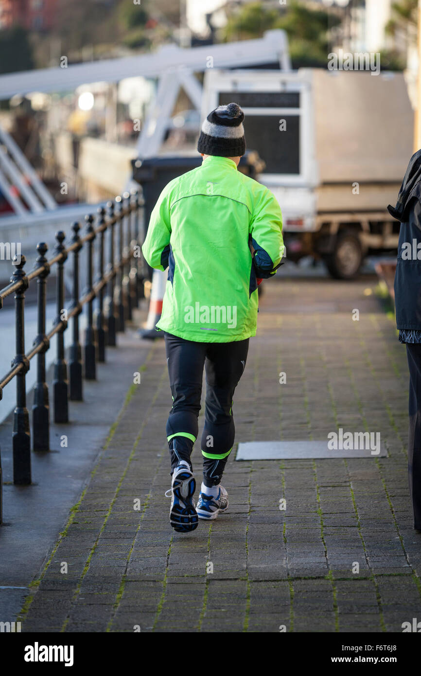 Ein älterer Mann trägt eine wollige Pudelmütze, läuft Trainingsanzug Hose und einem grünen hi-viz oben weg von der Kamera auf den Hafen. Stockfoto