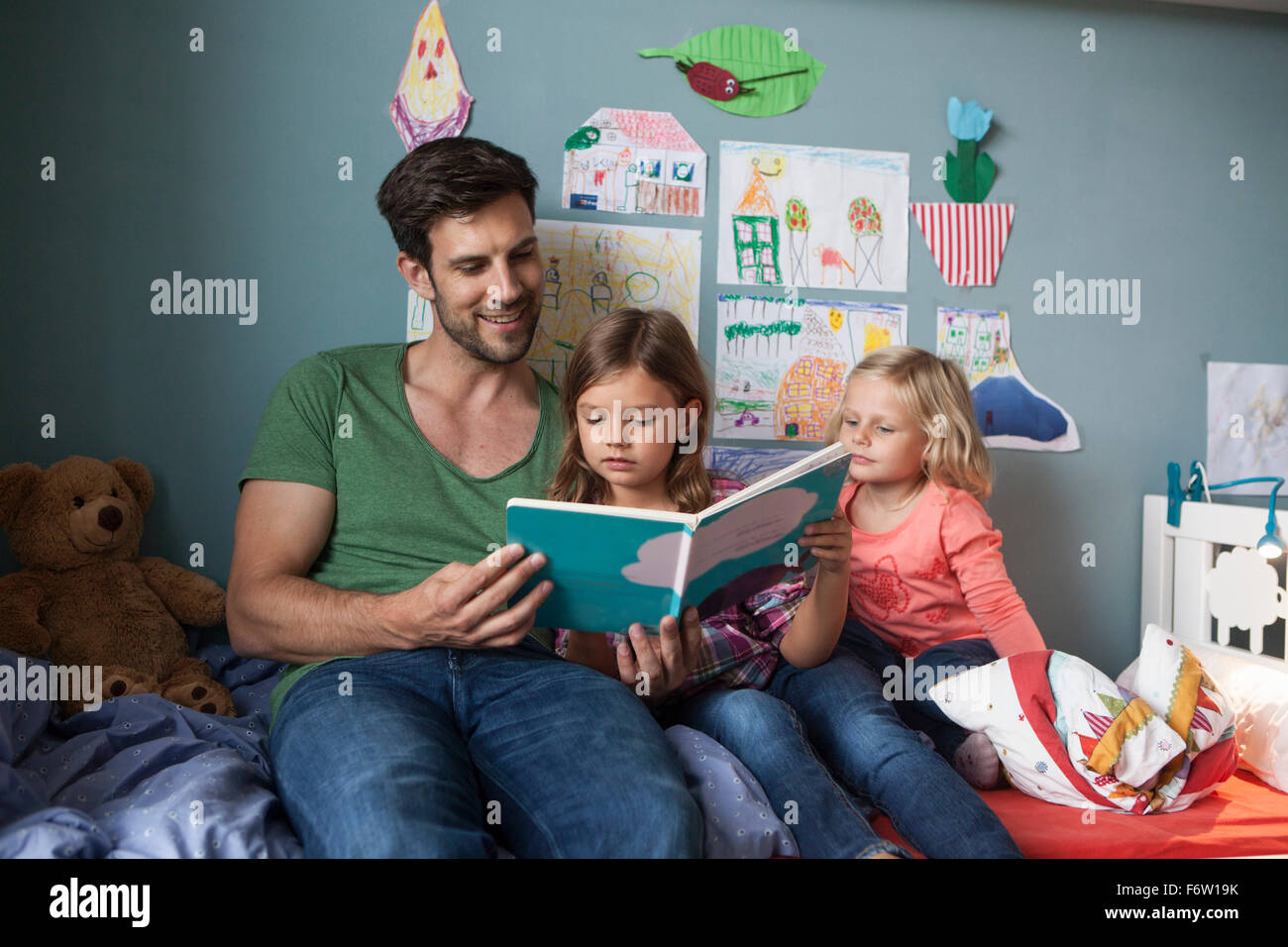 Vater und seine kleinen Töchter sitzen zusammen auf dem Bett im Kinderzimmer ein Buch zu lesen Stockfoto