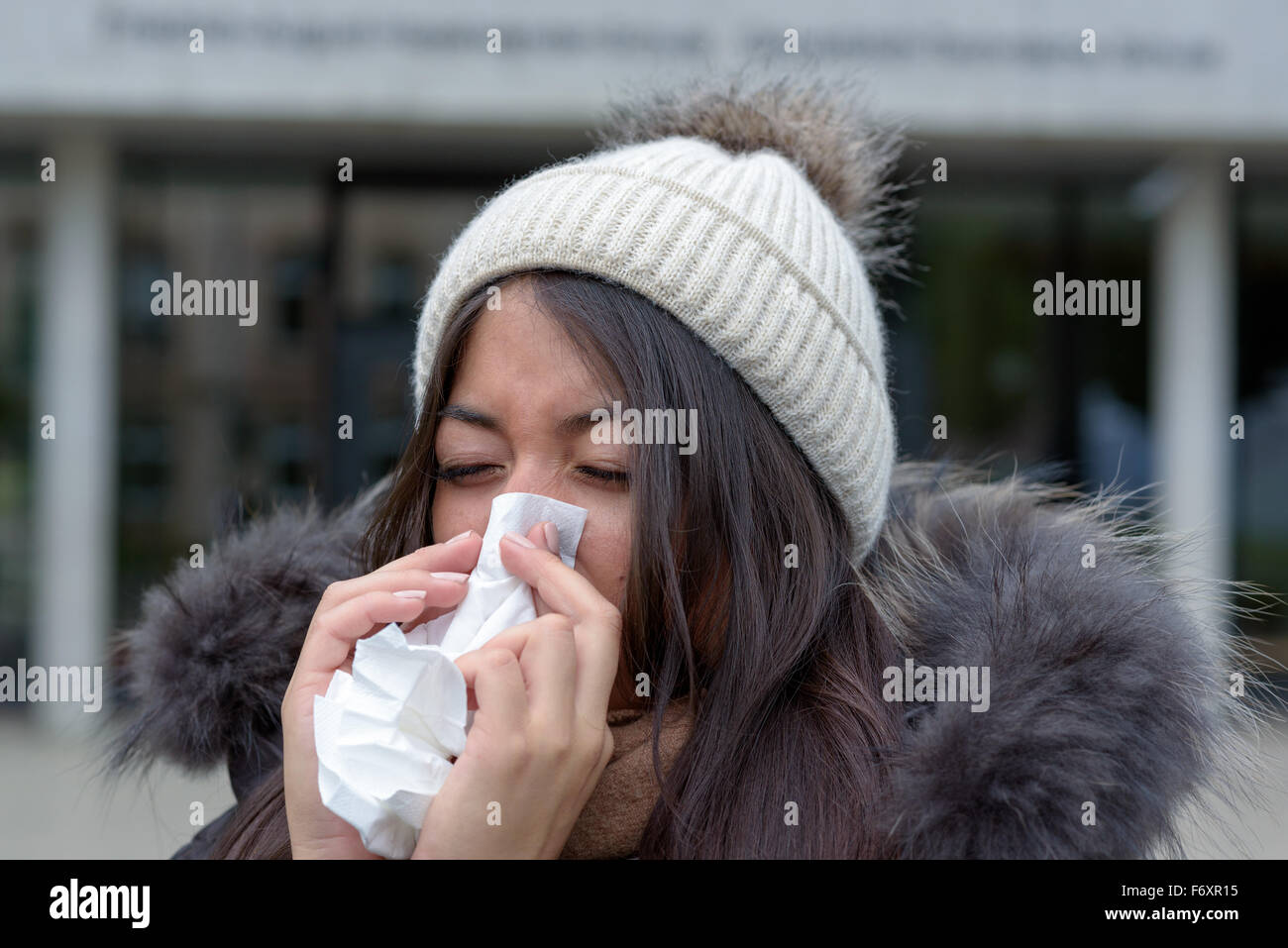 Junge Frau mit einer saisonalen Winterkälte oder Grippe trägt einen pelzigen Jacke und ihre Nase auf ein weißes Taschentuch o Strickmütze Stockfoto