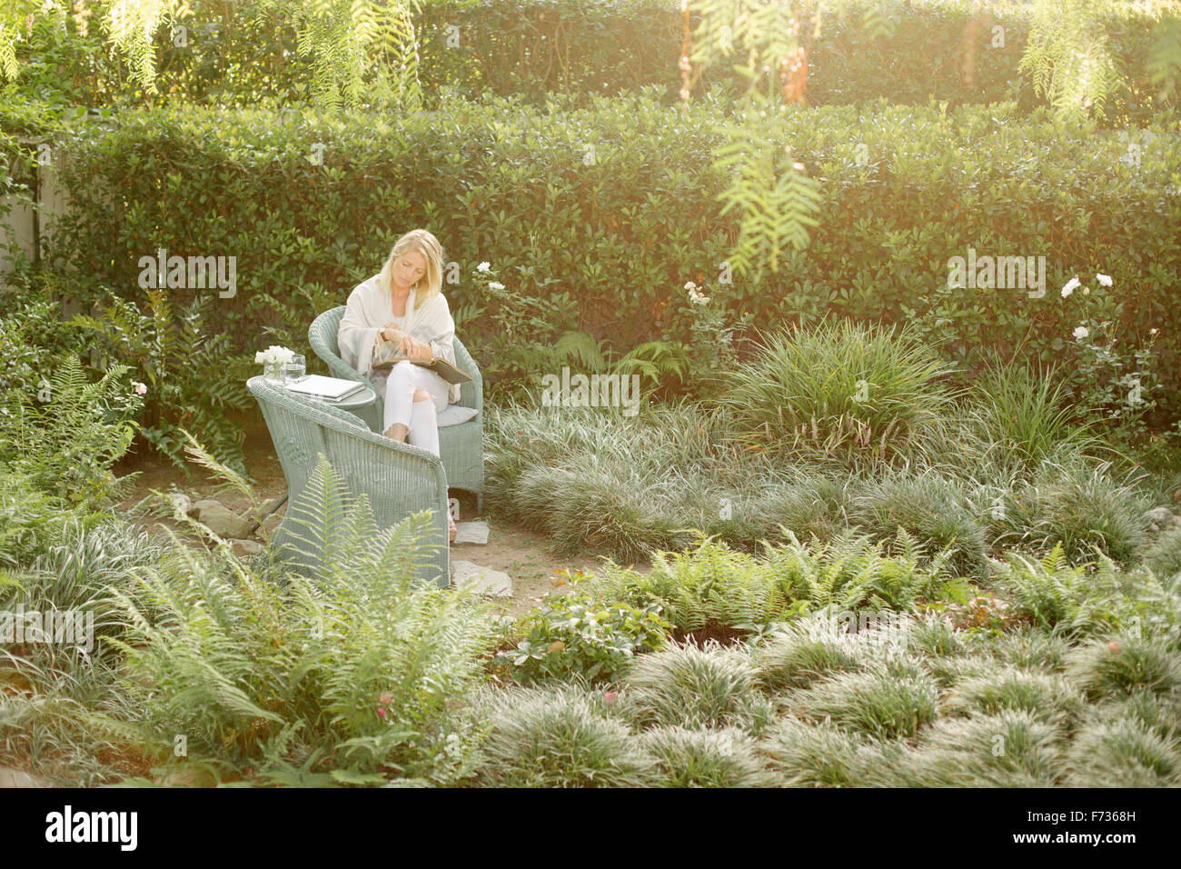 Blonde Frau sitzt in einem Korbstuhl in einem Garten lesen. Stockfoto