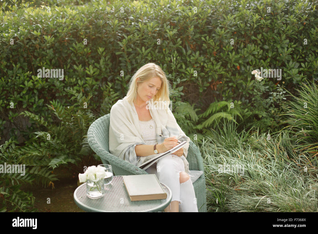 Blonde Frau sitzt in einem Korbstuhl in einem Garten, schreiben. Stockfoto