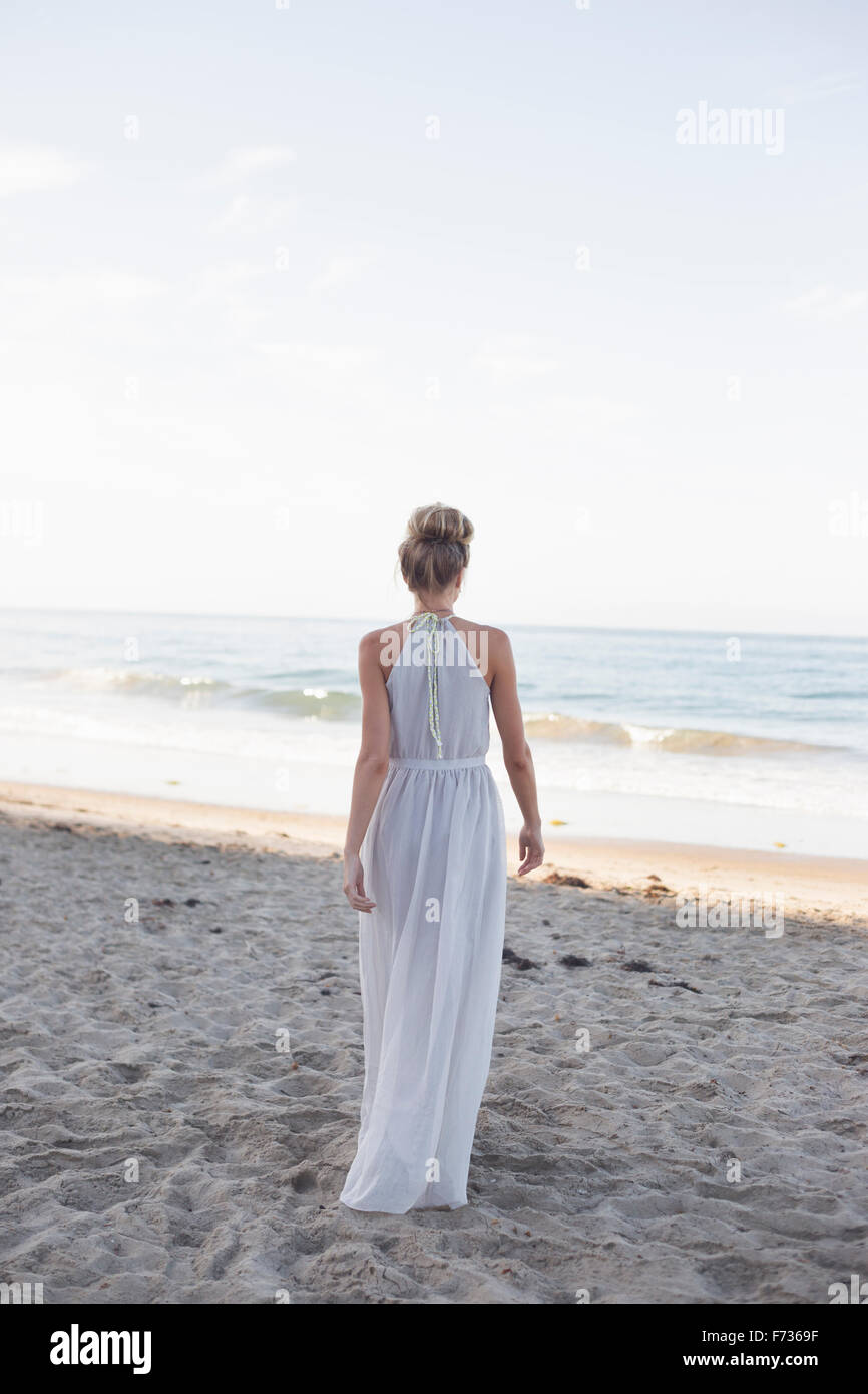 Blonde Frau trägt ein langes Kleid steht an einem Sandstrand. Stockfoto