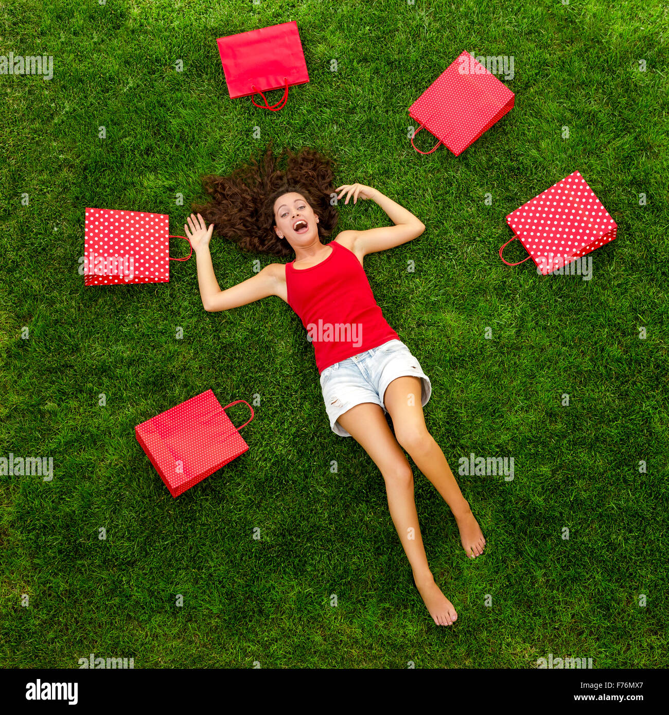 Schöne und glückliche junge Frau liegend auf dem Rasen, umgeben von roten Geschenk-Taschen Stockfoto