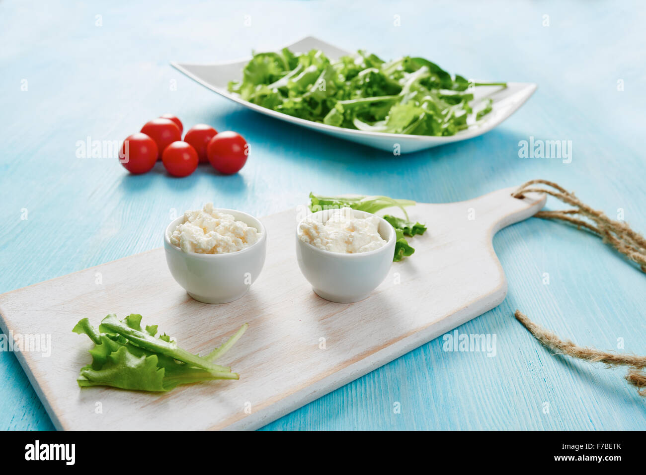 Hüttenkäse in zwei weißen Schalen auf blauen Holztisch, Salat und Tomate Stockfoto