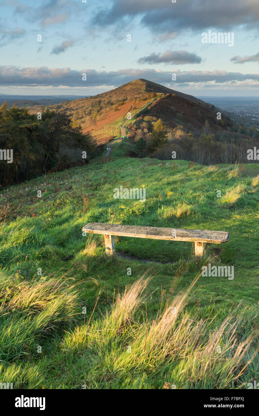 Eine Holzbank auf Jubillee Hügel von wunderschönen Sonnenuntergang, der auch Beleuchtung Ausdauer Hügel hinauf und Worcestershire Beacon ist beleuchtet Stockfoto