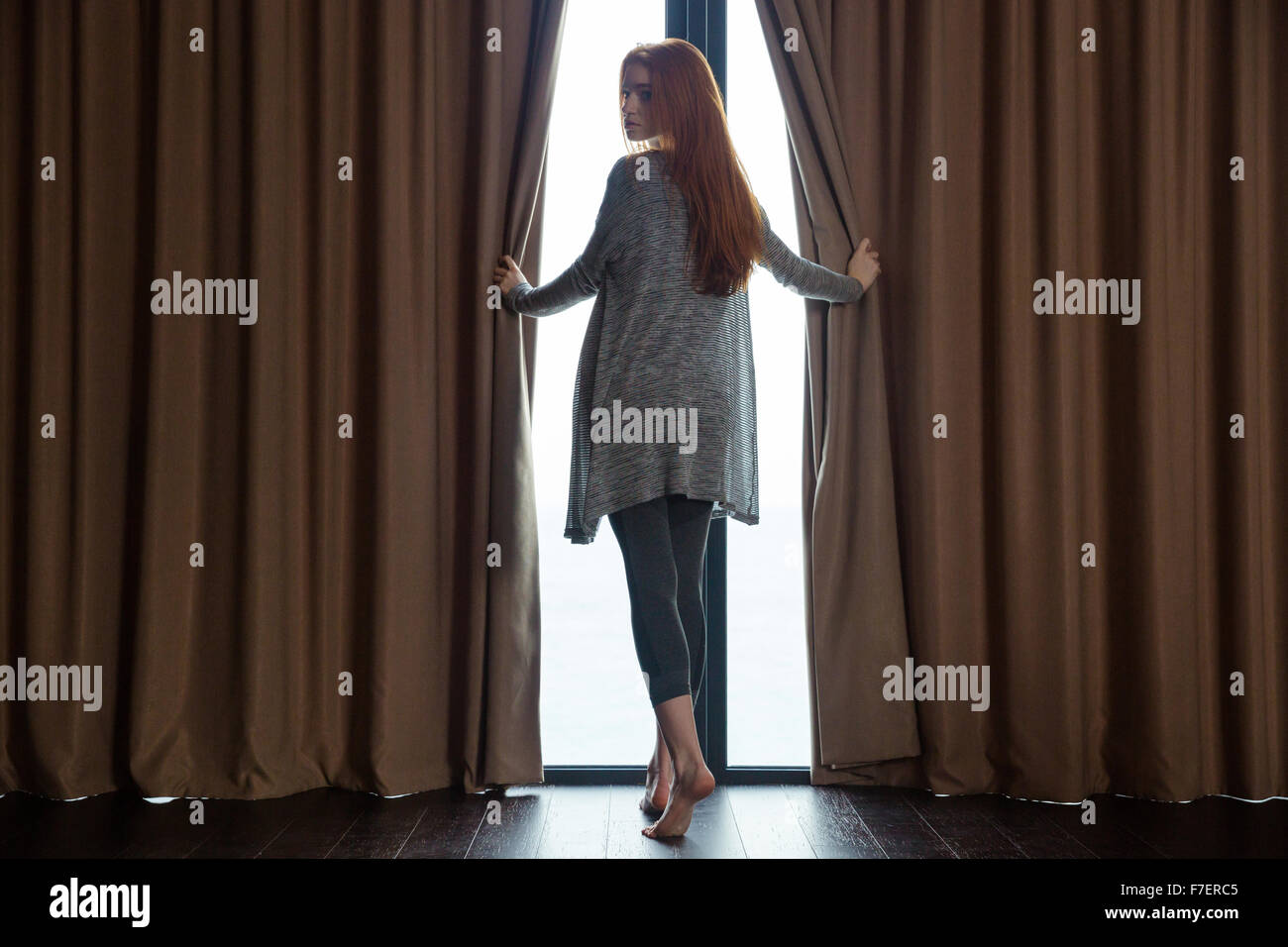 Entspannte schöne rothaarige junge Frau öffnen der Vorhänge, barfuß vor dem Fenster stehen und blickte zurück Stockfoto