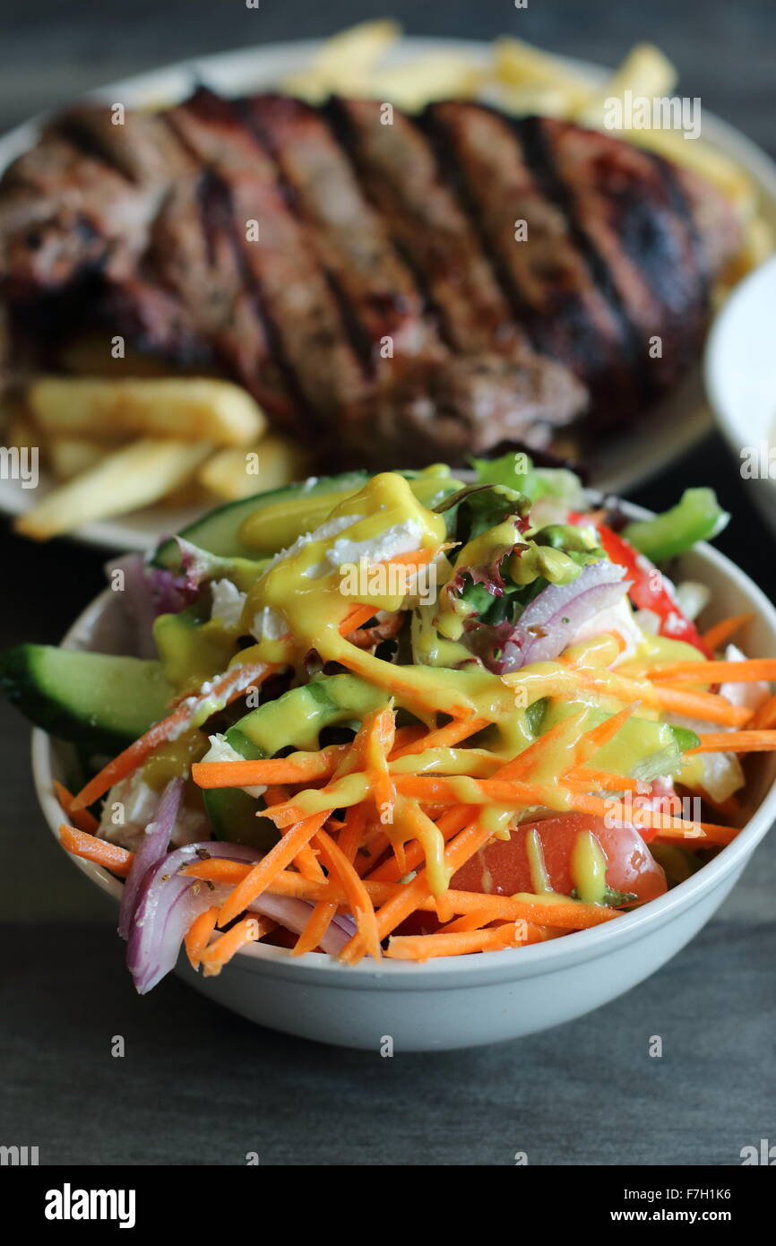 Eine Schüssel mit griechischer Salat in eine weiße Schüssel mit Steak und Chips im Hintergrund Stockfoto