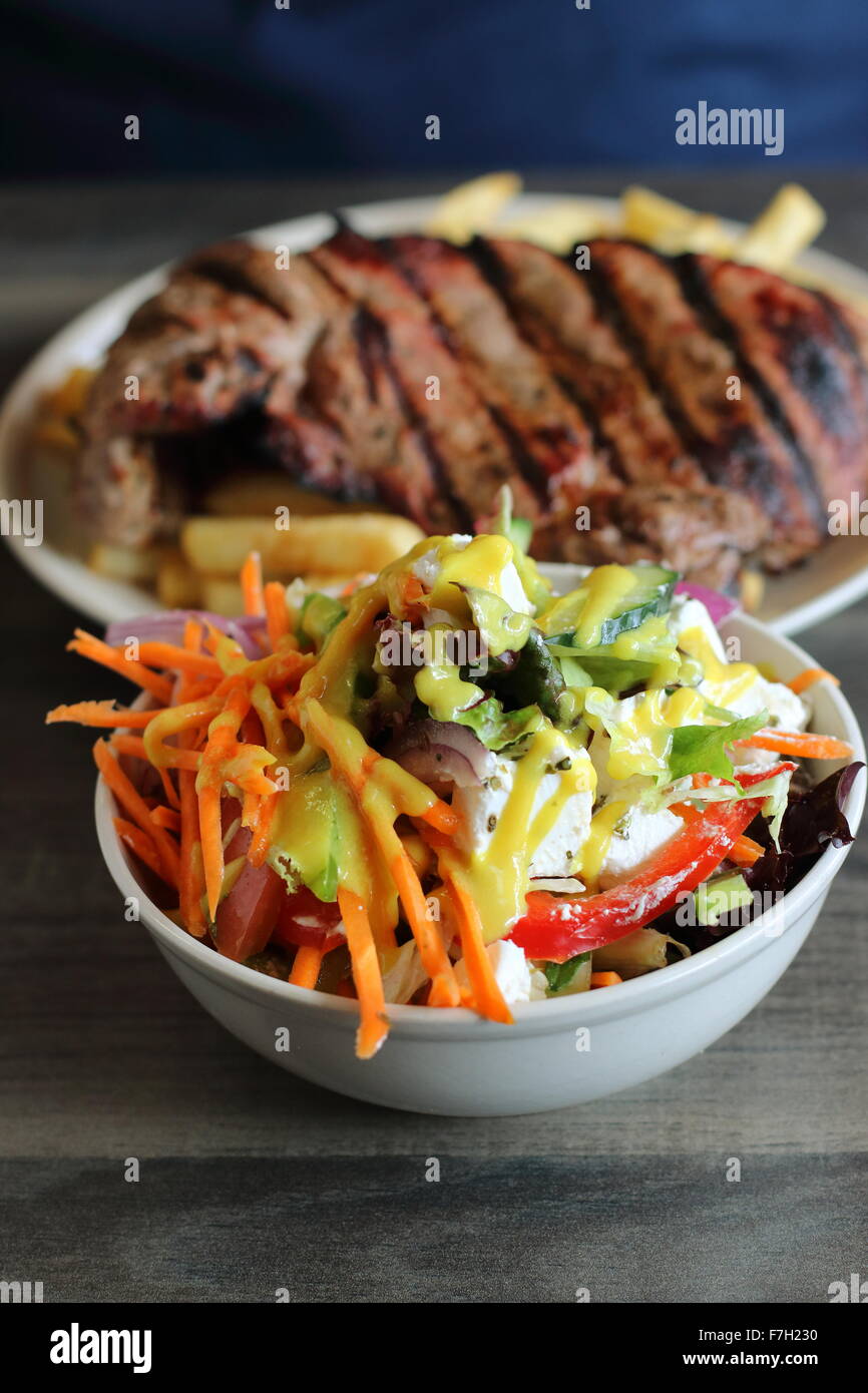 Eine Schüssel mit griechischer Salat in eine weiße Schüssel mit Steak und Chips im Hintergrund Stockfoto