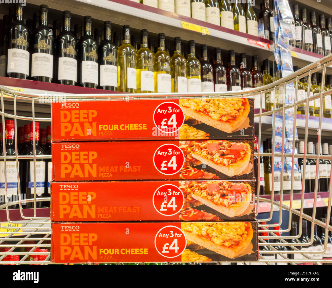 Vier gefrorene Tesco Deep Pan Pizzas im Einkaufswagen mit Wein im Hintergrund. Gesundes/ungesundes Diät-Konzept... Tesco Store, Großbritannien Stockfoto