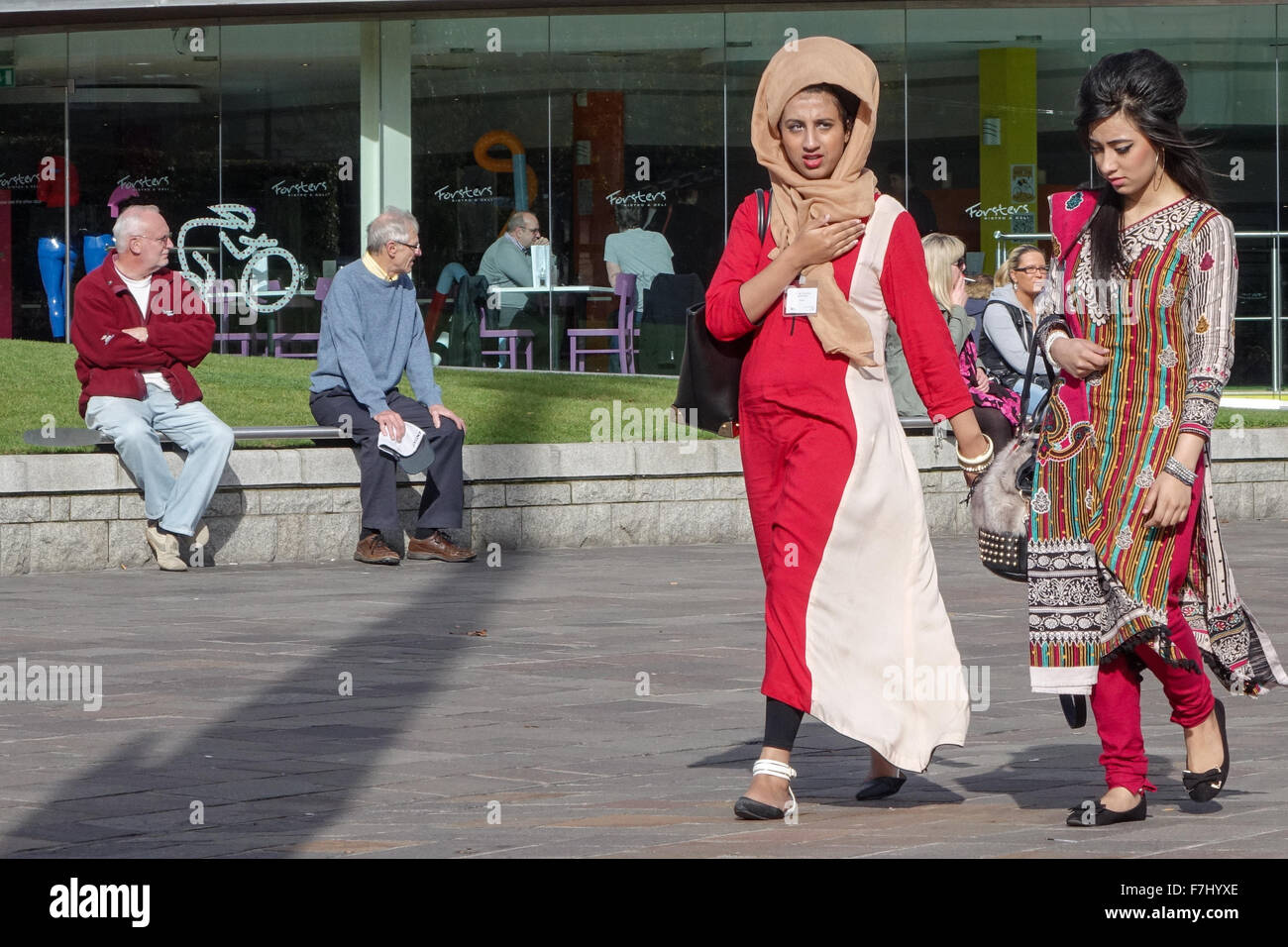 Zwei muslimische Frauen, die auf dem Bradford Centenary Square spazieren, tragen traditionelle östliche Kleidung mit zwei älteren Männern im Hintergrund Stockfoto