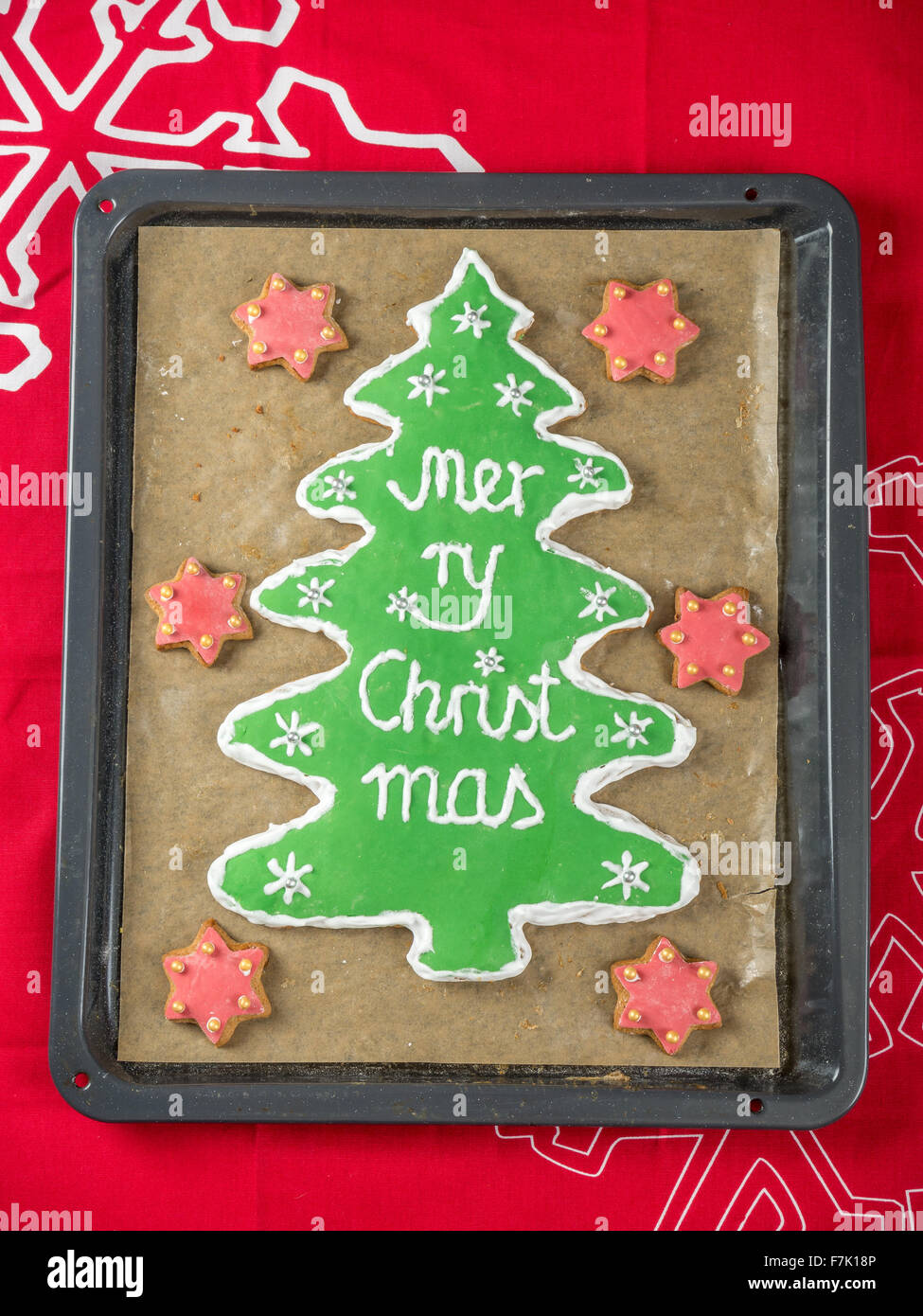 Hausgemachte Weihnachtsbaum-wie Lebkuchen mit grüner Glasur und Frohe Weihnachten schreiben auf Backblech gelegt am roten Tisch Stockfoto