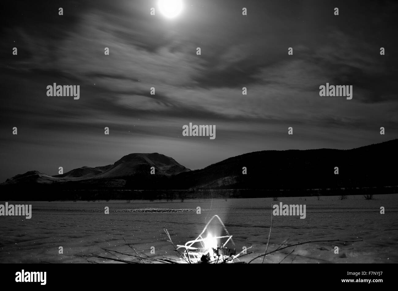 brennendes Feuer auf gefrorenen Flussbett mit majestätischen schneebedeckten Berg und Vollmond im Hintergrund Stockfoto