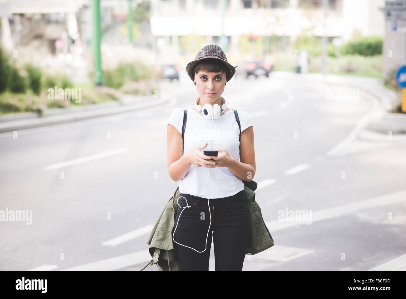 Knie Figur des jungen schönen kaukasischen braun glattes Haar Frau posiert in der Stadt, die eine Smartphone Kopfhörer um den Hals, auf der Suche in der Kamera, heiter - Technologie, soziales Netzwerk, Kommunikations-Konzept Stockfoto