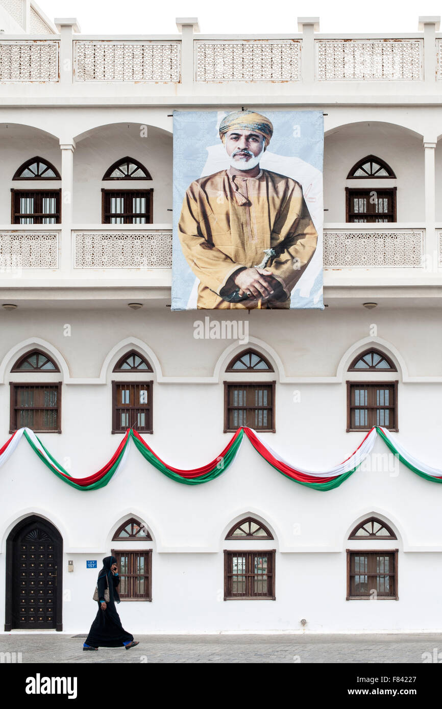 Frau zu Fuß vorbei an einem Gebäude geschmückt mit einem Plakat des Sultans von Oman in Mutrah in Muscat, der Hauptstadt des Oman. Stockfoto
