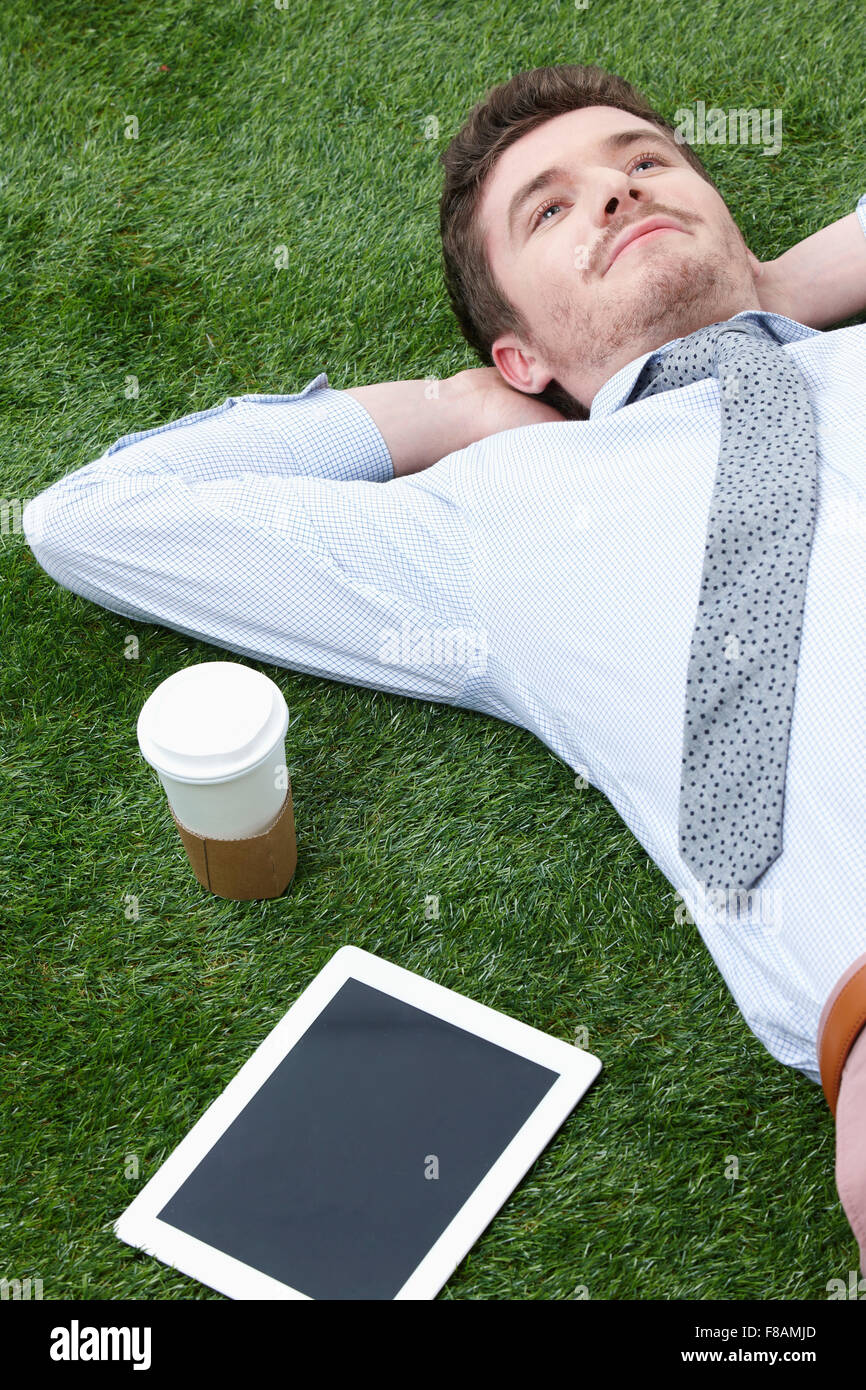 Porträt des Denkens Geschäftsmann liegend mit dem Kopf auf die Arme auf Grünland mit Take away Kaffee und tablet Stockfoto