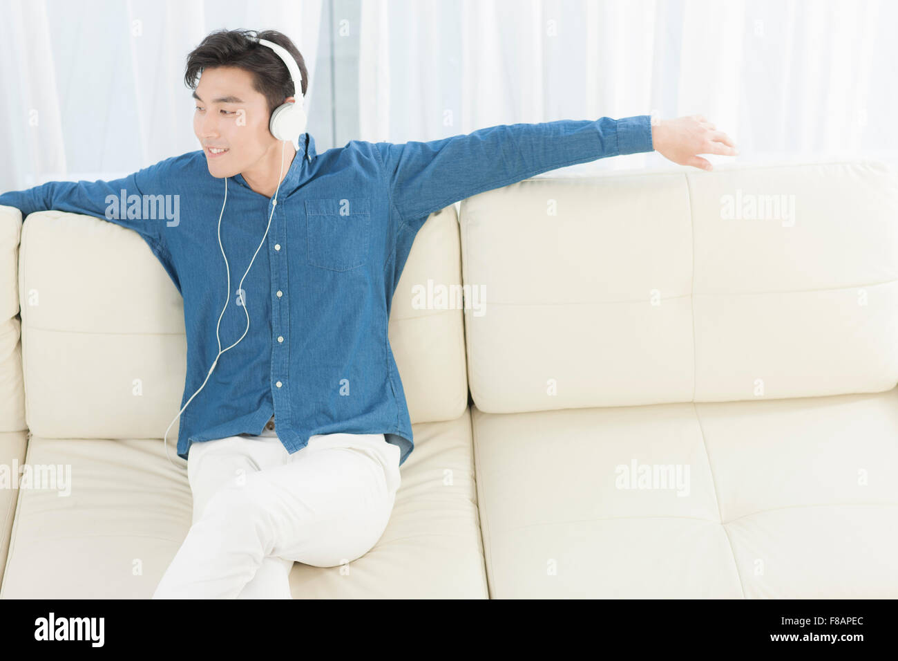 Lächelnd jungen Mann auf Sofa mit gekreuzten Beinen sitzen Musikhören Arme öffnen Stockfoto