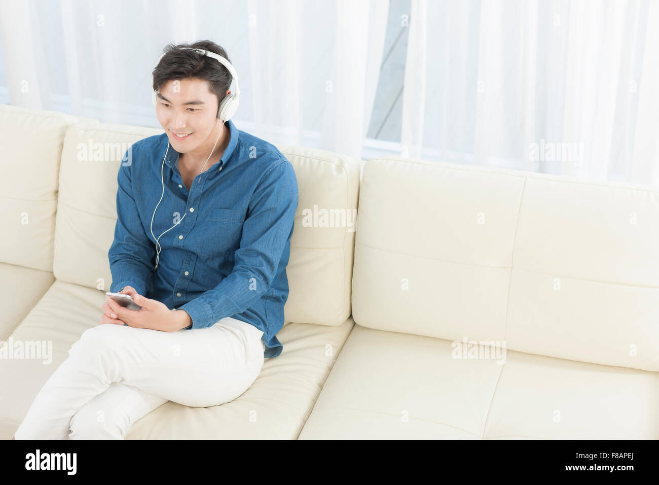 Lächelnd jungen Mann auf Sofa sitzen mit gekreuzten Beinen Musikhören auf smartphone Stockfoto