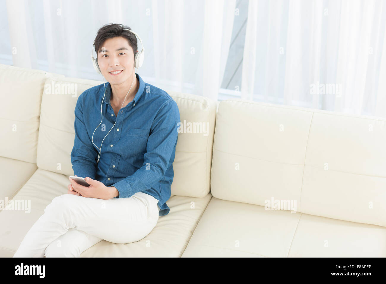 Lächelnd jungen Mann auf Sofa sitzen mit gekreuzten Beinen Musikhören auf Smartphone starrte auf Vorderseite Stockfoto