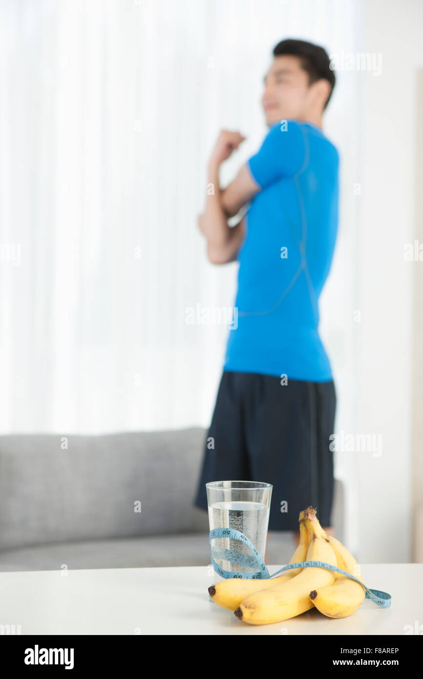 Junger Mann stehend stretching mit Wasserglas, Bananen und Maßband auf Tisch Stockfoto