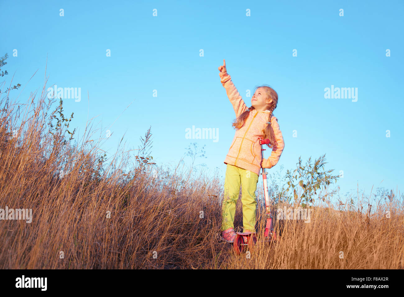 Kleine Mädchen in helle Kleidung mit Scooter zeigt einen Finger in den Himmel auf die Natur Stockfoto