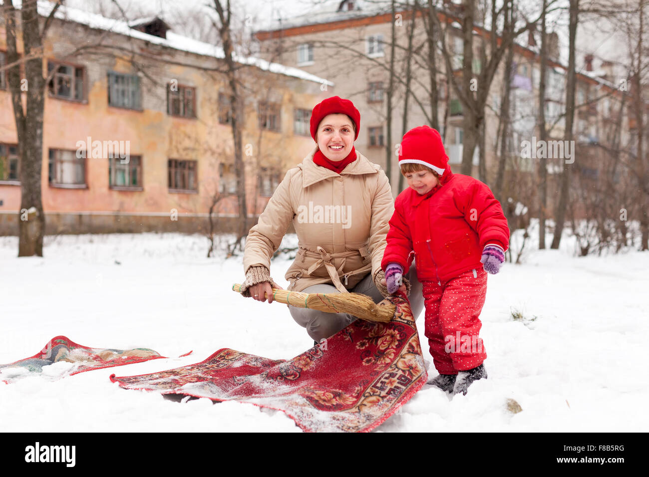Lächelnde Mutter mit Kind reinigt Teppich mit Schnee im Wintertag im freien Stockfoto