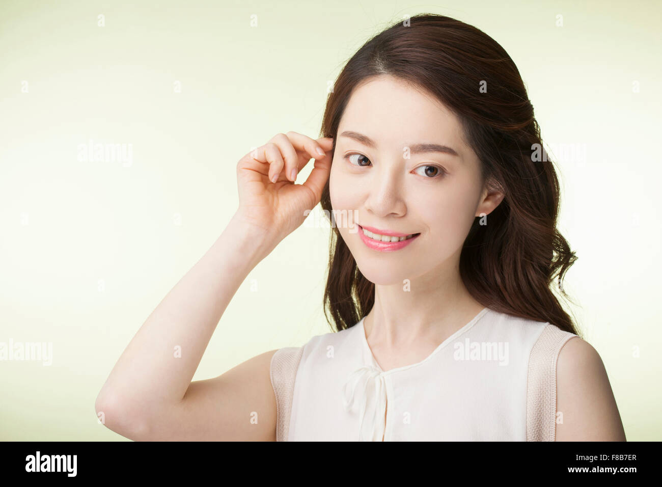 Koreanerin mit lange gewellte Haare in weißen ärmellosen Shirt ihr Haar zurück in ihr Ohr zu ziehen und auf der Suche zur Seite mit einem Lächeln Stockfoto