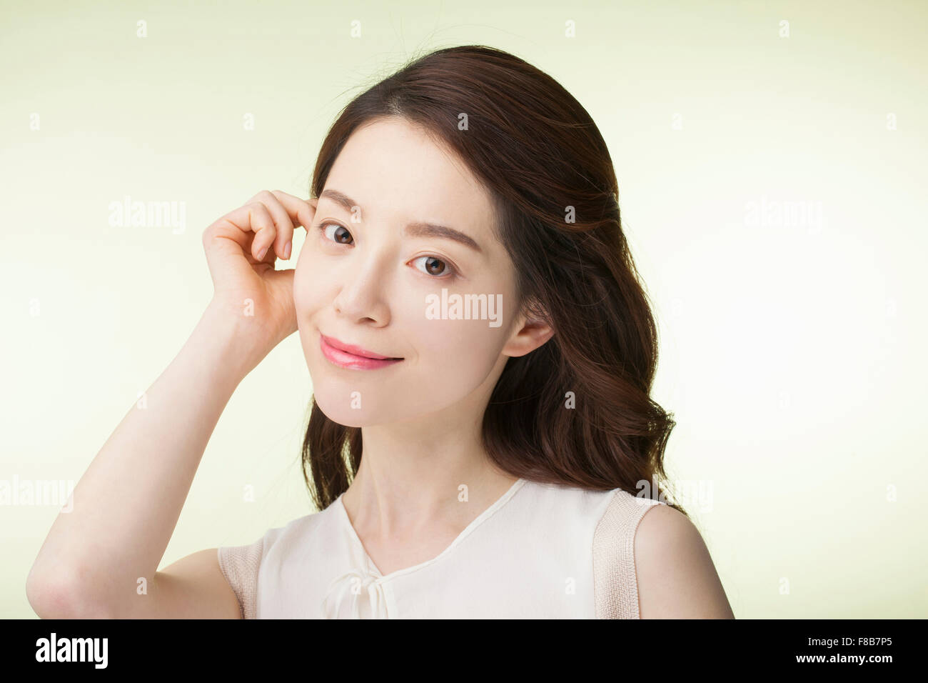 Koreanerin mit lange gewellte Haare in weißen ärmellosen Hemd zeichnen ihr Haar zurück in ihr Ohr und lächelnd nach vorne Stockfoto