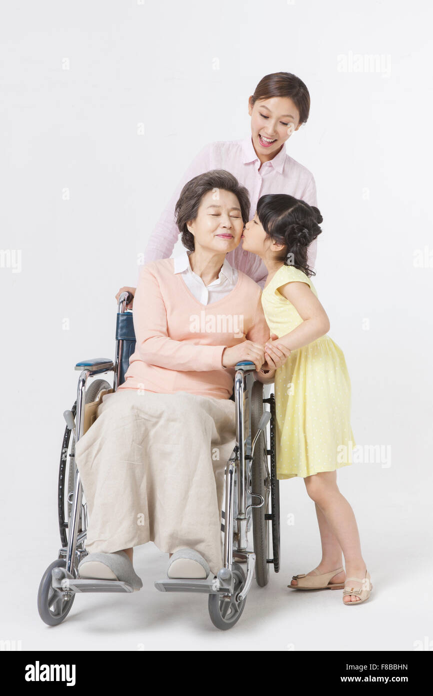Mutter sitzt im Rollstuhl und Tochter halten die Griffe und küssen zu ihrer Großmutter Enkelin Stockfoto