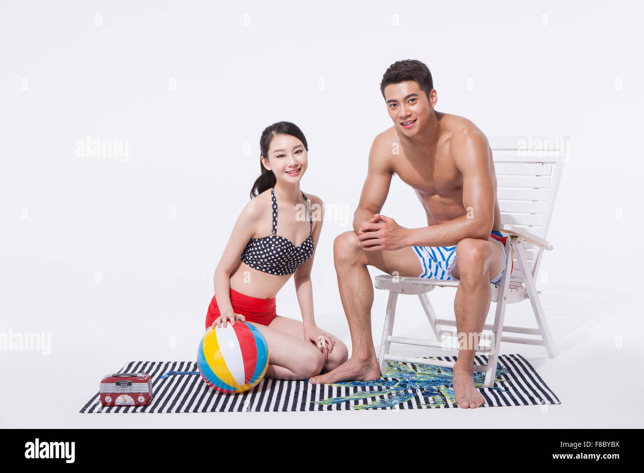 Frau kniete auf einem Handtuch mit einem Wasserball und ein Mann sitzt auf einem Stuhl in Badebekleidung starrte mich mit einem Lächeln Stockfoto Frau kniete auf einem Handtuch mit einem Wasserball und ein Mann sitzt auf einem Stuhl in Badebekleidung starrte mich mit einem Lächeln Stockfoto