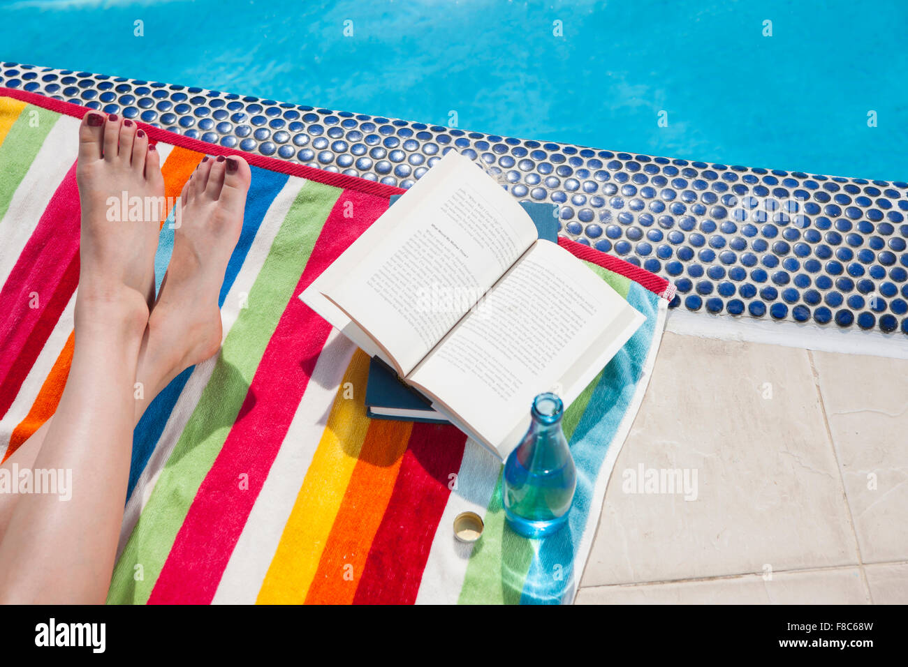 Hohen Winkel der gekreuzten Beinen einer Person auf einem bunten Tuch mit Büchern und Getränken am Rande eines Swimmingpools Stockfoto