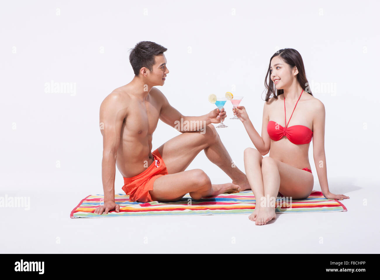 Paar in Strand zu tragen, sitzt auf einem Badetuch und Toasten mit Cocktails einander beide sahen einander Stockfoto