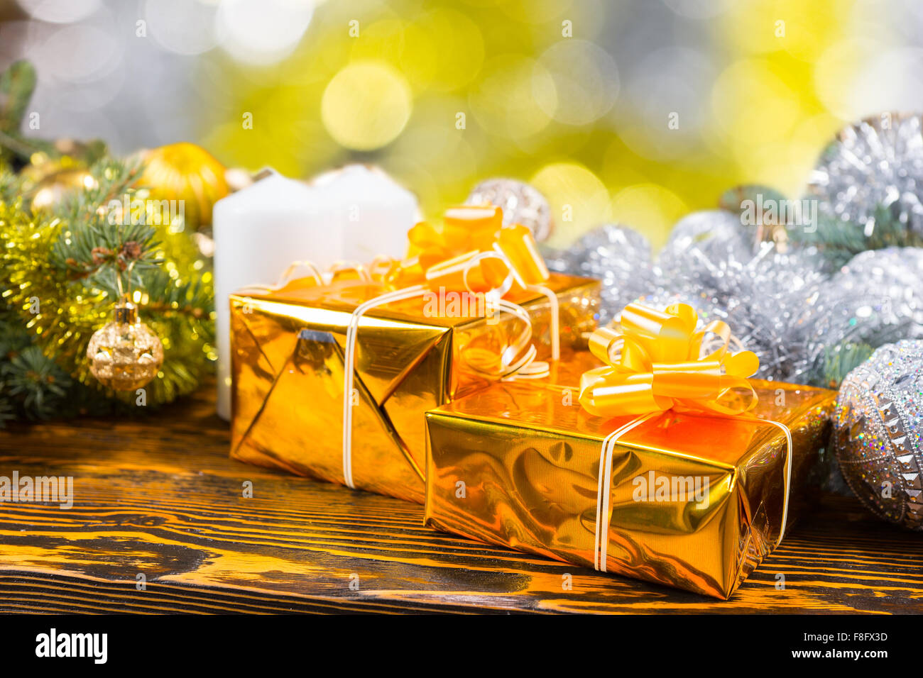 Nahaufnahme von zwei Geschenke verpackt in Gold mit Bögen neben weißen Stumpenkerzen auf rustikalen Holztisch, umgeben von immergrünen Zweigen mit Lametta Garland und Kugeln geschmückt. Stockfoto