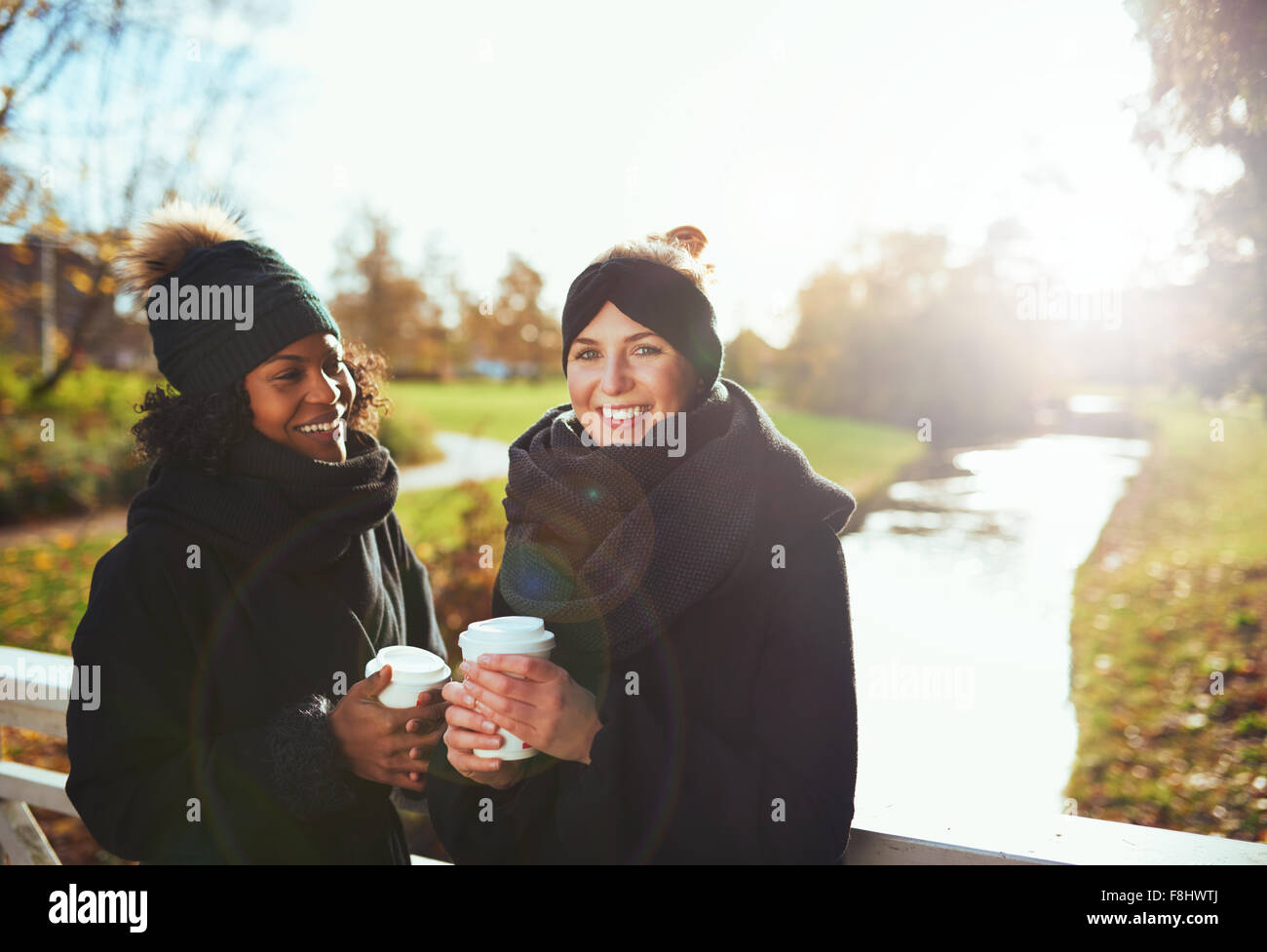 Zwei Freundinnen Blick in die Kamera und lächelnd stehen auf Brücke, halten Kaffee zum mitnehmen, Fluss im Hintergrund Stockfoto