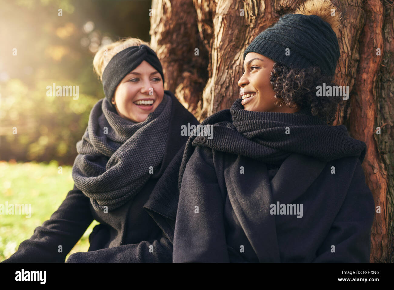 Zwei junge Frauen stützte sich auf Baumstamm im herbstlichen Park und sahen einander lächelnd in Nahaufnahme Stockfoto