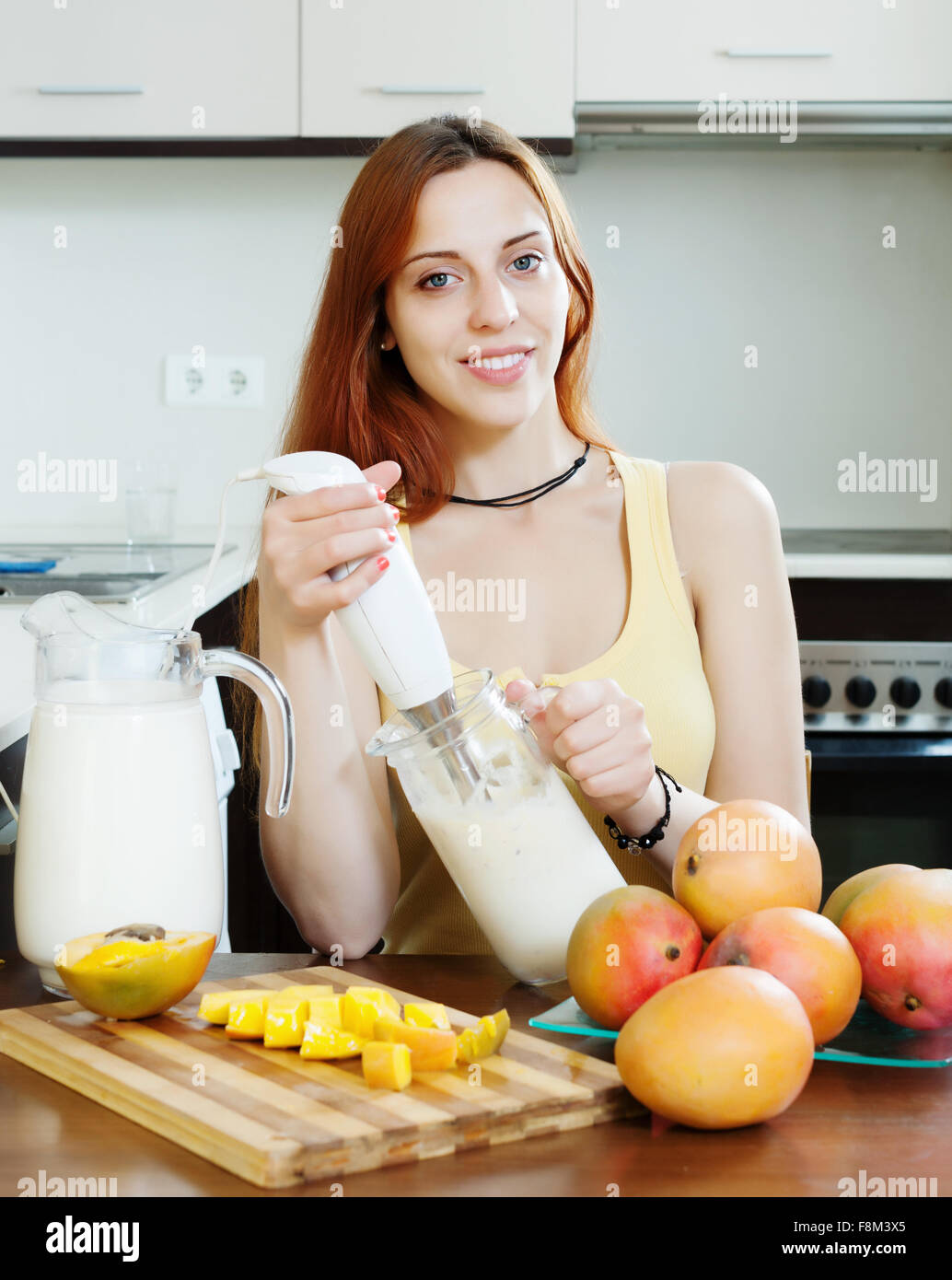 gewöhnliche junge Frau, die Zubereitung von Getränken mit Blender von Mango und Milch in der heimischen Küche Stockfoto