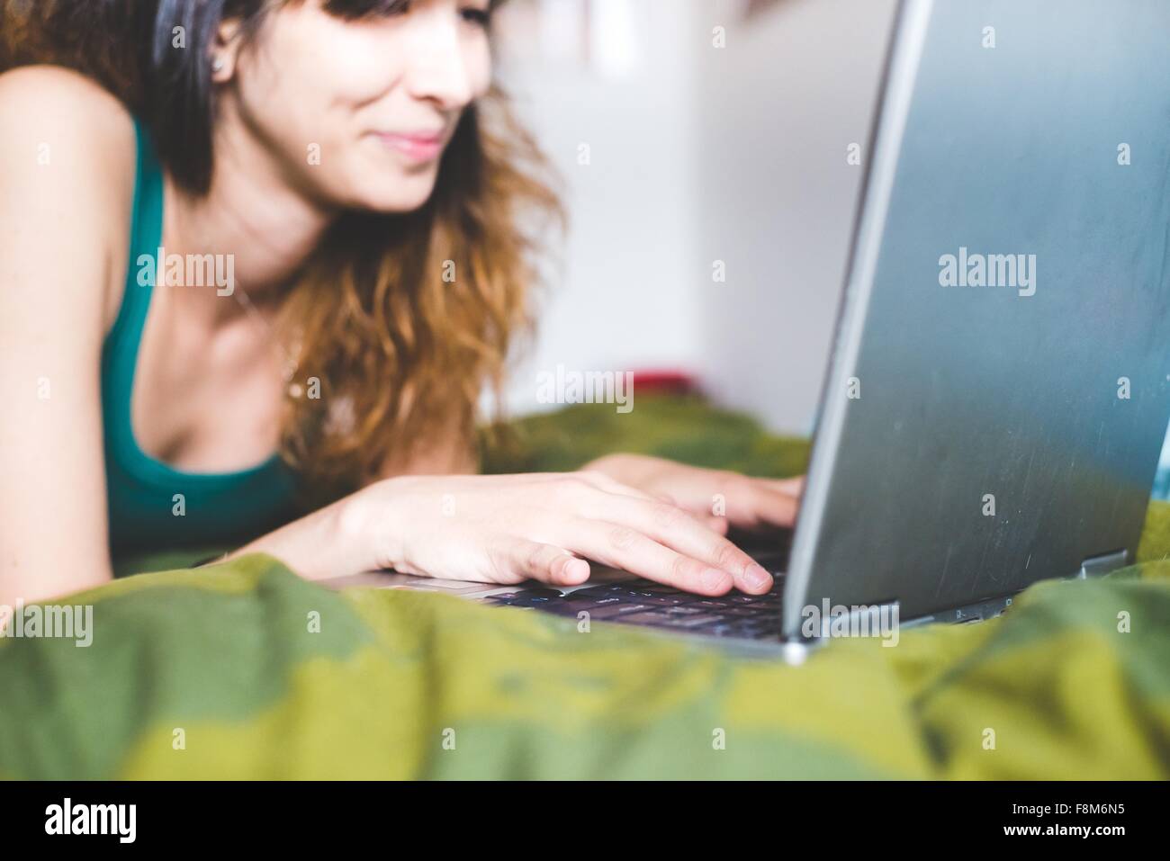 Junge Frau liegend auf Vorderseite auf Bett mit Laptop Computer lächelnd, differenzielle Fokus Stockfoto