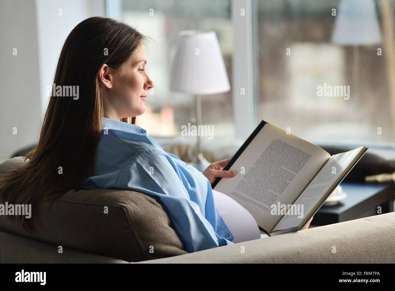Schwangere Frau auf Sofa sitzen, Buch lesen Stockfoto