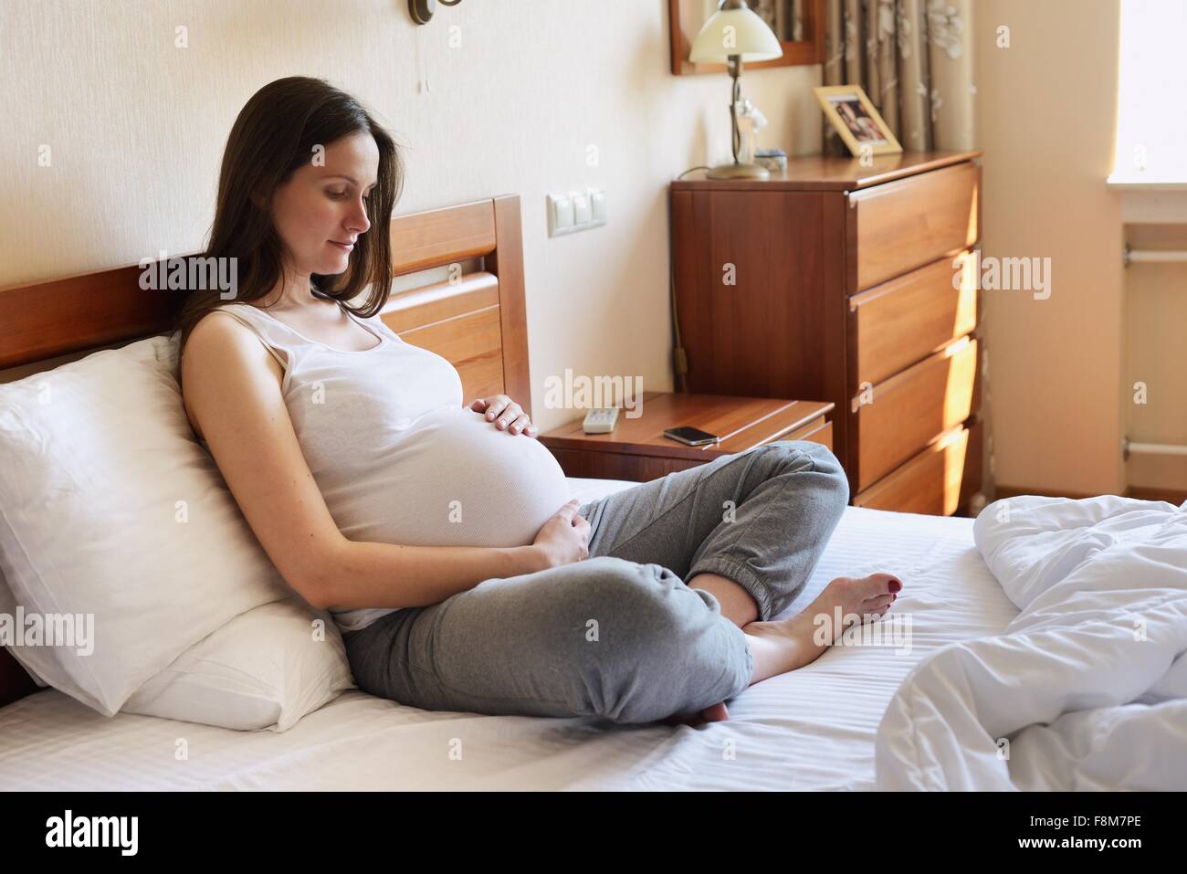 Schwangere Frau sitzt auf dem Bett, hält Magen Stockfoto